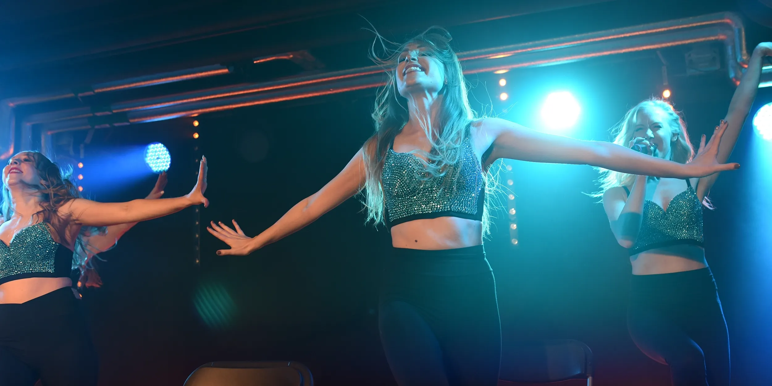 Tre showartister uppträder i synkron koreografi med utsträckta armar under en energifylld scenföreställning.