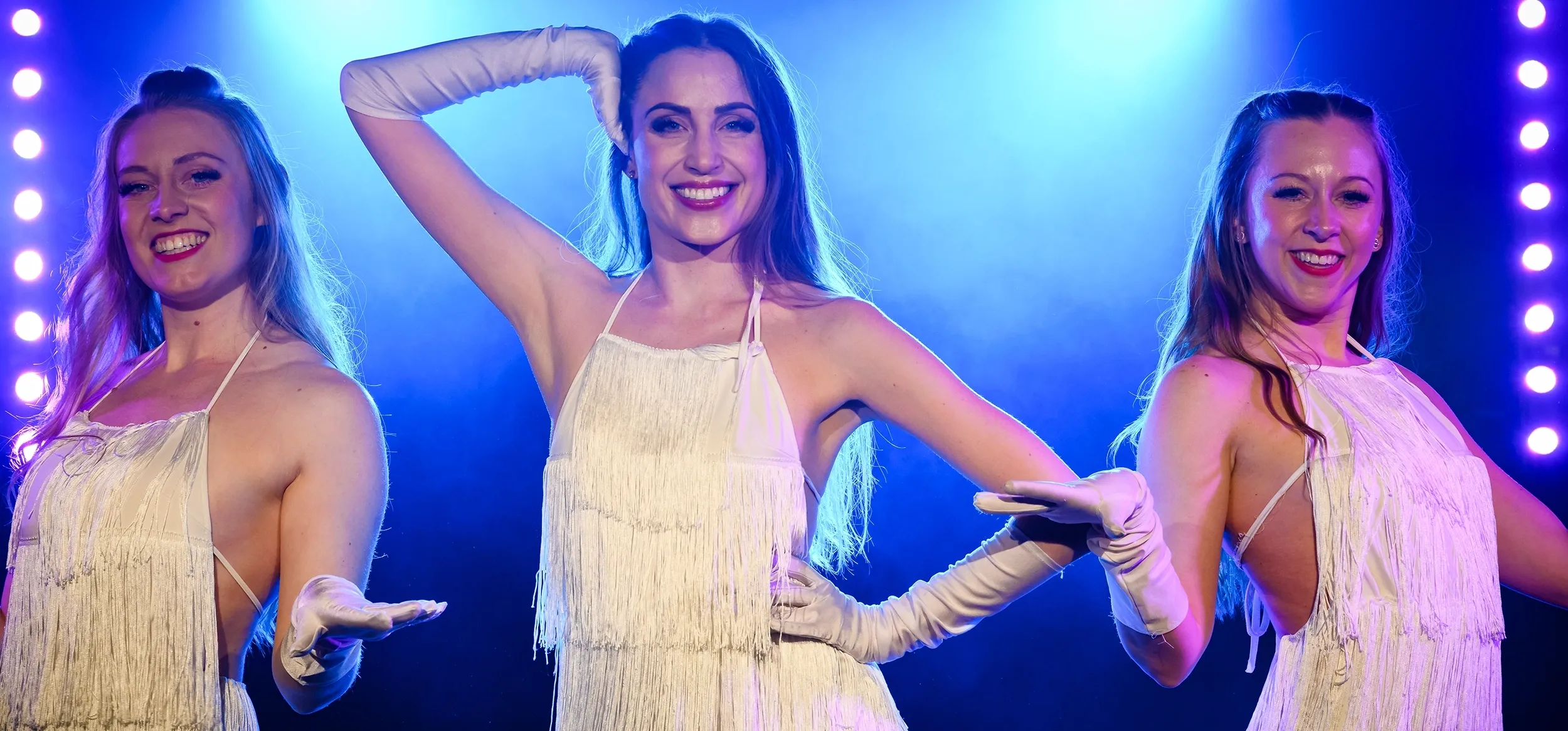 Tre showartister i ljusa scenkostymer poserar leende på scen i blått ljus under en professionell show.