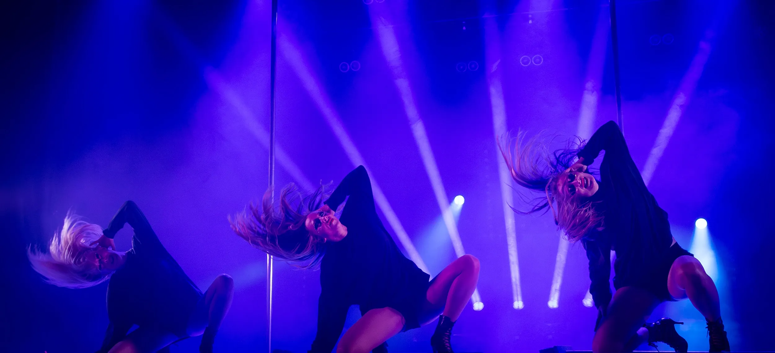 Tre dansare på scen i dramatisk blå och lila ljussättning, mitt i en kraftfull koreografisk rörelse med hår i rörelse och starkt scenuttryck.